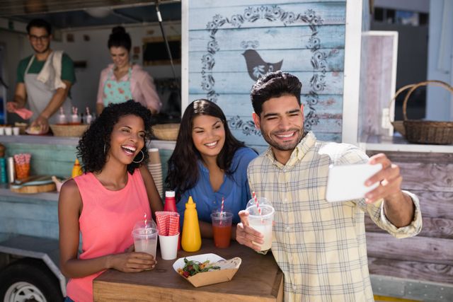 Group of friends enjoying time together at a food truck while taking a selfie. Perfect for promoting social gatherings, food truck businesses, and summer activities. Great for use in marketing materials, blogs, and social media posts related to outdoor dining and friendships.