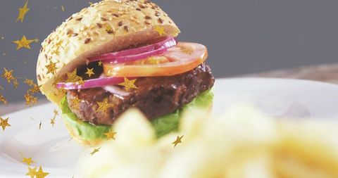 Sesame Seed Burger Sitting on White Plate with Gold Star Confetti and Blurred Fries