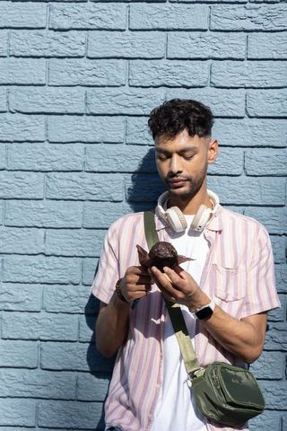 Indian man inspecting chocolate muffin while wearing headphones by blue brick wall