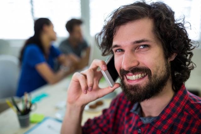 Male graphic designer with curly hair and beard talking on mobile phone in a modern office. He is smiling and appears to be in a casual and creative work environment. Colleagues are seen in the background collaborating. Ideal for use in articles about creative professions, teamwork, modern workspaces, and communication technology.