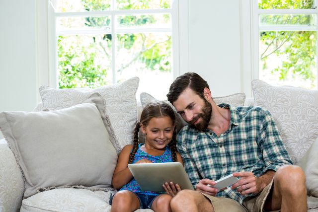 Happy girl showing digital tablet to father sitting on sofa