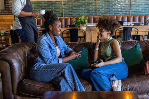 African american women chatting on leather sofa in cozy urban coffeehouse with smartphones