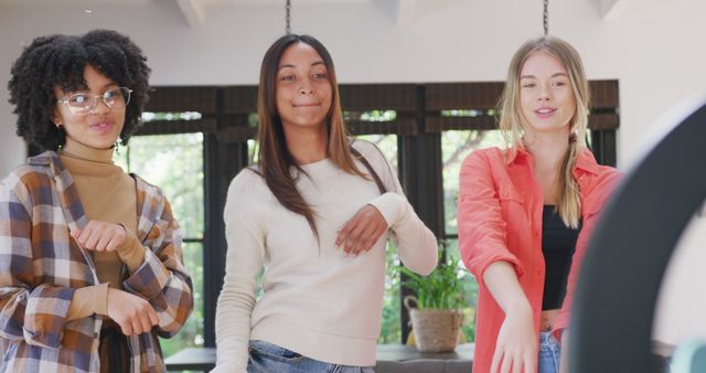 Three diverse young women are dancing while recording a video indoors. They are having fun, smiling, and appear to be filming content for social media. This image is perfect for use in articles, advertisements, and social media posts related to friendship, social media trends, youth culture, and entertainment.