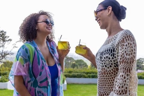 African american women clinking refreshing yellow cocktails on patio in colorful coverups