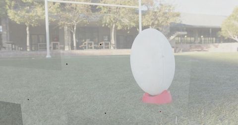 Modern white oval sculpture resting on pink base on grassy campus with goalpost background