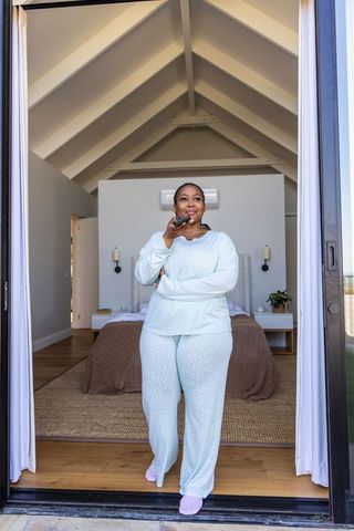 African american woman standing at bedroom doorway in blue pajamas using smartphone