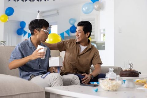 Asian father and son celebrating birthday at home on couch opening gifts and smiling