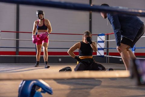 Female boxer sitting on ring canvas holding face recovering from knockdown while coach assisting