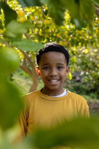 Smiling african american boy standing in sunlit orchard wearing mustard shirt among green fruit