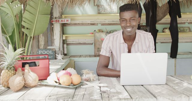 Portrait of happy african american man using laptop in surf shop on beach. Lifestyle, business, vacation, summer and leisure, unaltered.