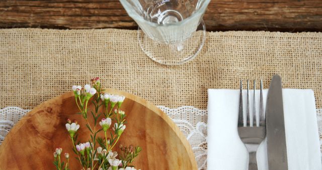 A rustic table setting features a wooden plate with a small bouquet of flowers, silverware wrapped in a white napkin, and an elegant glass, with copy space. The arrangement suggests a warm, inviting atmosphere for a meal in a country or vintage-themed setting.