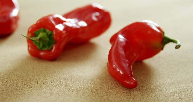 Three red chili peppers are placed on a beige surface, showcasing their vibrant color and fresh appearance. Their glossy texture and curved shapes suggest they are ripe and spicy, ideal for culinary uses that require a kick of heat.