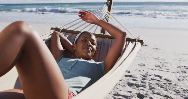 Young woman lounging in a hammock on a sandy beach with the ocean in the background, enjoying a sunny summer day. Perfect for concepts related to vacations, leisure time, relaxation, outdoor activities, and carefree lifestyle.