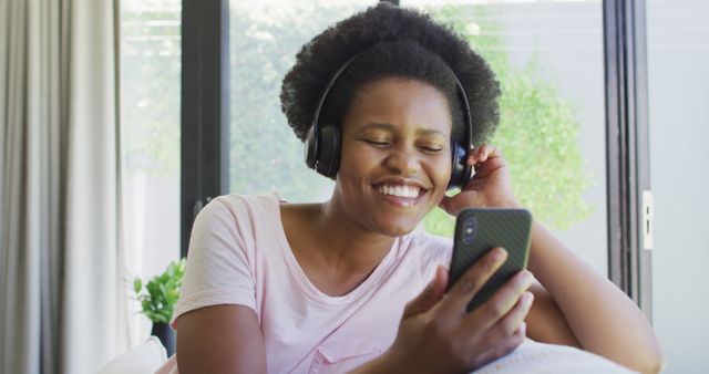 Young African American woman using headphones and smartphone while sitting indoors, smiling and enjoying music. Ideal for illustrating concepts related to technology, leisure time, happiness, and the use of modern devices.