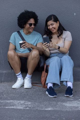Laughing diverse friends sitting on curb sharing phone and coffee in urban casual scene