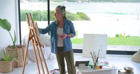 Mature woman painting at easel in sunlit coastal studio holding drink and brushes