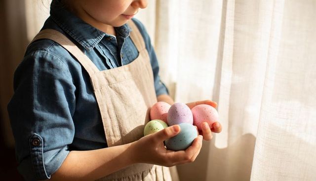 Sunlit child holding pastel speckled easter eggs wearing denim shirt and linen apron