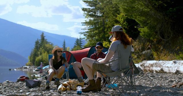 Friends are sitting in camping chairs by a mountain lake, with a tent nearby. Use this for outdoor adventure promotions, travel blogs, or friendship-themed content.