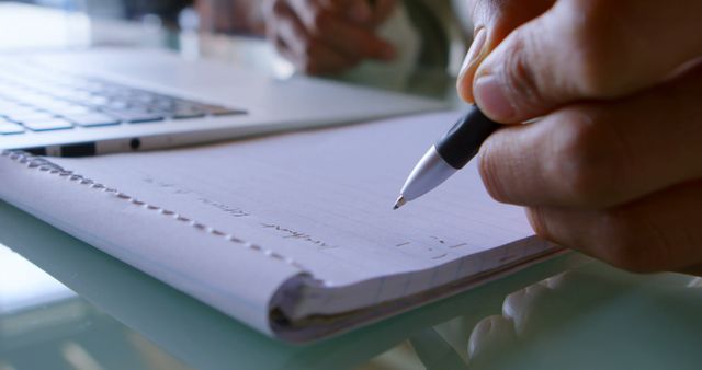 Close-up of person's hand writing on a notepad with a pen beside a laptop. Could be used for portraying themes of studying, working, note-taking, remote work, or office environment.