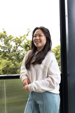 Young asian woman standing on balcony smiling in cream hoodie and light blue sweatpants