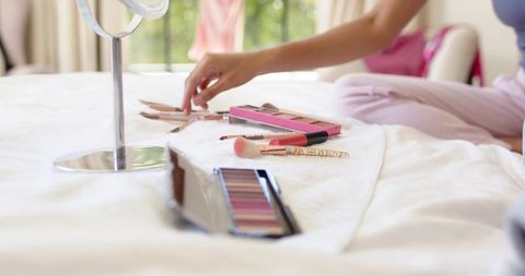 Woman in pink pajamas reaching for eyeshadow palette on bed morning beauty routine