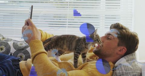 Young man cuddling kitten on sofa wearing mustard sweater, holding phone, cozy daylight