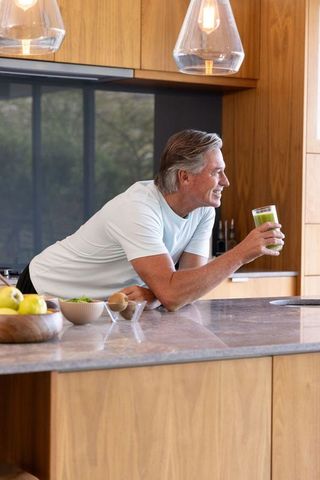 Smiling senior man leaning on modern kitchen island holding green smoothie