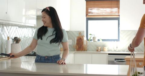Asian woman cooking and chatting with partner in sunlit contemporary kitchen