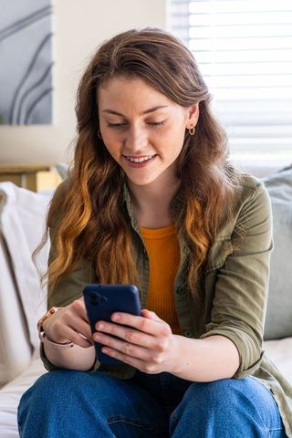 Young woman sitting on sofa texting on smartphone in relaxed at-home scene with sunlight