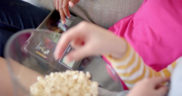 This playful moment captures the essence of modern digital entertainment for children. Two youngsters are seen enjoying their time with a streaming video on a tablet while indulging in a bowl of popcorn. Ideal for marketing campaigns, educational articles on children's technology use, or advertisements for streaming services and snack brands aimed at young audiences.