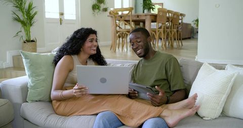 Diverse couple working from home on laptop and tablet while relaxing on sunlit modern sofa