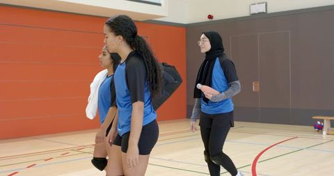 Multicultural female volleyball team walking across school gym carrying bags and towel