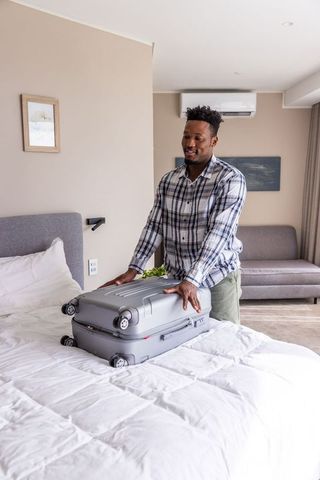 African american man packing gray hardshell suitcase on bed in minimalist guest room