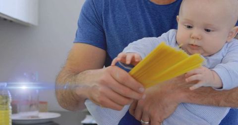 Father holding baby reaching for uncooked spaghetti in home kitchen, playful bonding moment