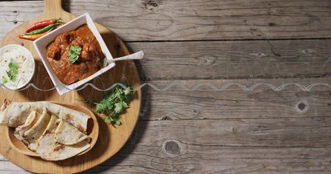 Serving spicy curry with flatbreads and yogurt on round board overhead flatlay, cilantro