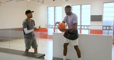 Diverse male friends holding basketball and carrying gym bag on indoor court concourse