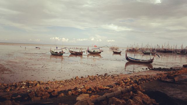 Fishing boats resting on the shore during low tide under a cloudy sky. Use image for travel blogs, coastal tourism promotion, or websites about traditional maritime activities. Perfect for illustrating peaceful seaside locations or the challenges of coastal fishing.