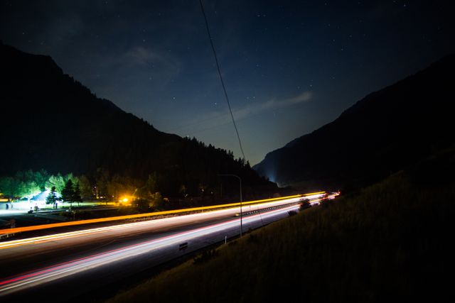 This night scene captures light trails from vehicles traveling on a mountain highway, surrounded by dark silhouetted mountains and under an expansive starry sky. The long exposure creates vibrant streaks of light and emphasizes the tranquility of the mountainous landscape. Ideal for travel blogs, articles on night photography, and promotional materials related to road trips and night-time adventures.