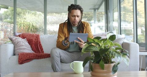 Mid adult man with dreadlocks using tablet on sofa in minimalist glass living room