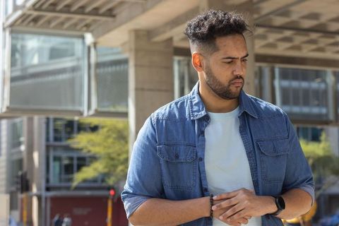Young man standing under urban overpass clasping hands, contemplative in denim shirt and smartwatch