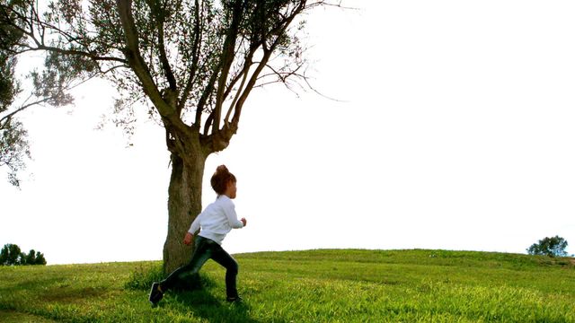 Child running playfully near solitary tree on grassy hill, capturing essence of freedom and joy. Useful for themes about childhood, outdoor play, nature, activity, or mental wellness concepts.