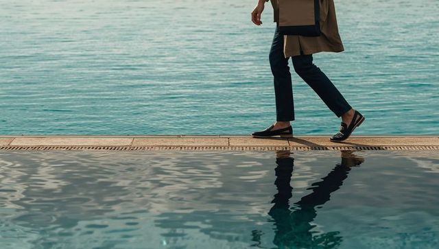 Walking in trench coat and loafers along waterfront pool with reflection and copy space