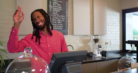 Smiling barista waving behind cafe counter wearing pink shirt using touchscreen pos