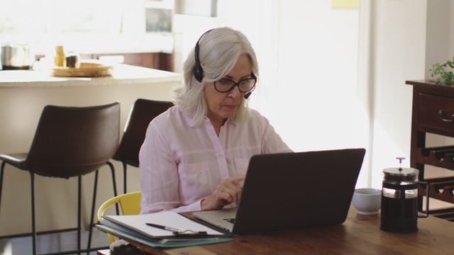 A senior woman sits at a table in a well-lit kitchen area, using a laptop while wearing headphones. This demonstrates a modern tech-savvy approach as she engages in remote work or online communication. Suitable for advertising telework, older adults embracing technology, or staying connected during challenging times such as a pandemic.