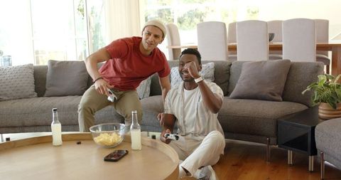 Diverse friends playing console game in bright living room, cheering over victory and snacks