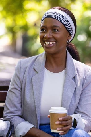 Smiling african woman enjoying takeaway coffee on park bench wearing blazer and headband