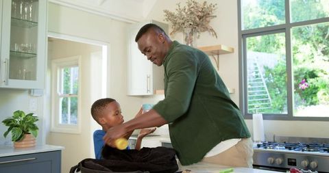 African american father and son packing backpack together in sunlit modern kitchen