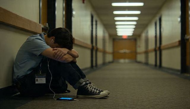 Teen sitting in dim school corridor listening to music on smartphone, conveying isolation