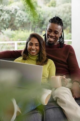 South asian woman and african american man relaxing on couch using laptop and sharing coffee