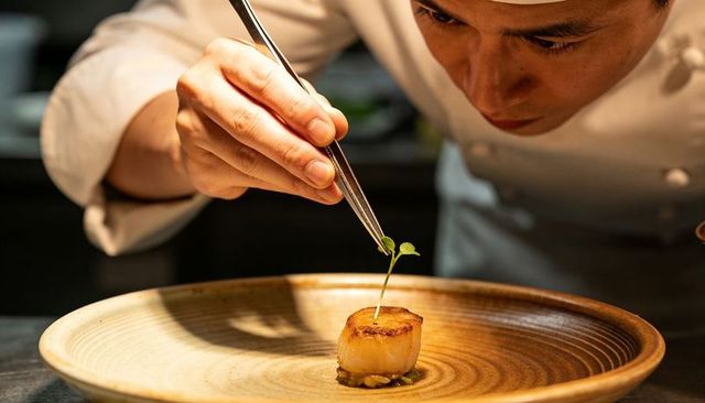 Chef placing microgreen on seared scallop with tweezers, fine dining plating artistry